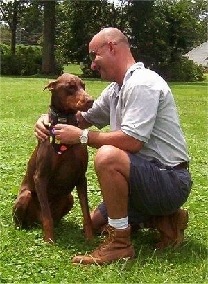 Boomer pruunikaspruunikas ja punakaspruun Doberman Pinscher istub põllul koerale naeratava kiilaspäise mehe kõrval, kellel on päikeseprillid Joe. Koer kannab musta bandanat, mille kohal on täpid.