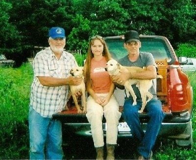 Tre personer på baksidan av en röd plockbil med en öppen bakgrind. Två män med en dam i mitten. Männen har basebollkepsar och blå jeans och håller solbruna och vita medelstora jakthundar.