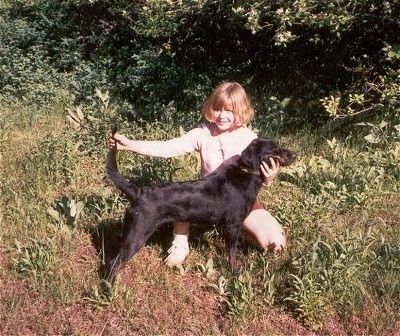 Rusvaplaukė mergina klaupiasi už „Leopard Cur“ šuns, pozuojančio jį kamino lauke, už jų - miškai.